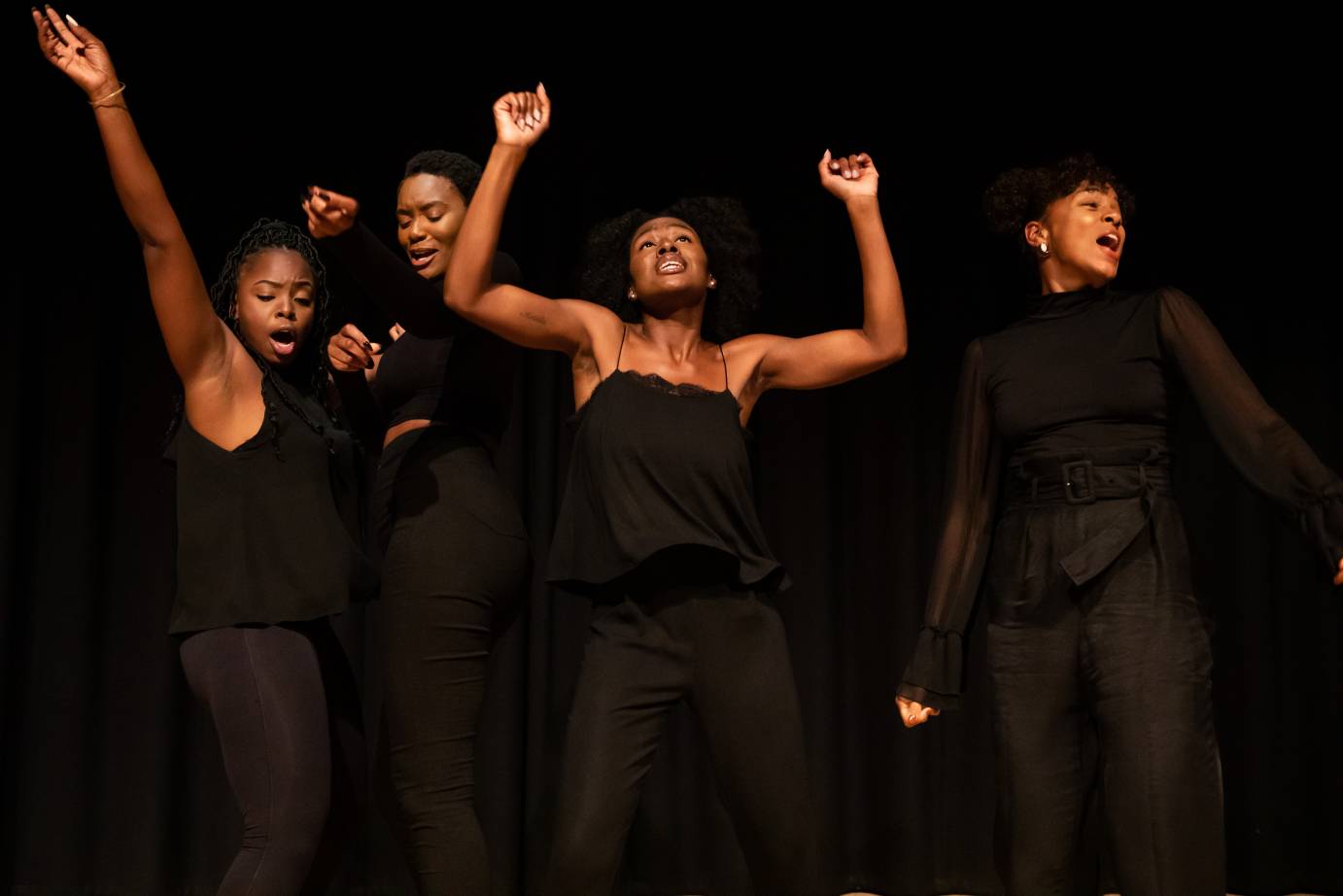 Four Black women dressed in black costumes - over the shoulder, sleeveless, to the chin. Center woman both arms upraised with bent elbows facing the audience, woman on left right arm extended on diagonal, woman on right with head turned over left shoulder, fourth woman head tucked 